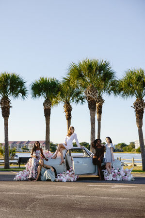 Five women posing around a pastel blue vintage convertible overflowing with pink and white flowers on a palm-lined waterfront road under a clear, sunlit sky