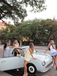 Five friends in summer dresses and sunglasses posing playfully around a light-blue vintage convertible parked on a historic southern oak-lined street draped with Spanish moss