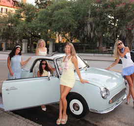 Five women in pastel summer dresses posing with a pale-blue vintage convertible on a tree-lined city street.