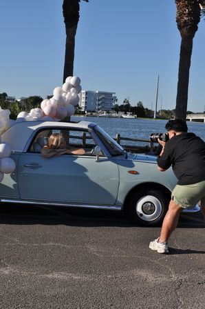Photographer shooting a model leaning from a pastel blue vintage convertible decorated with white and blush balloons at a palm-lined waterfront marina under a clear blue sky.