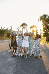 Bachelorette celebration: five women in sundresses pop champagne on a palm-lined sunset road beside a flower-covered car, champagne spray and raised arms