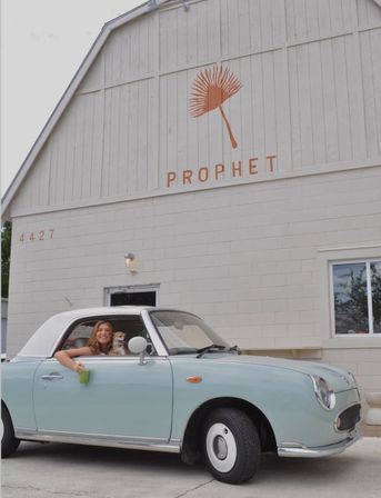 Woman leaning from the window of a pale blue vintage convertible, holding a green drink while a small dog rides shotgun in front of a cream-colored barn-style building with a stylized orange logo.