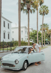 Romantic moment: a man kisses a woman’s hand while she sits in a pale blue vintage car on a palm-lined street in front of white historic houses.
