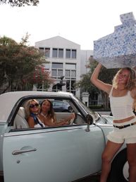 Smiling women posing with large floral-wrapped boxes beside a pale blue vintage convertible parked on a tree-lined downtown street in front of a white office building with pink flowering trees.