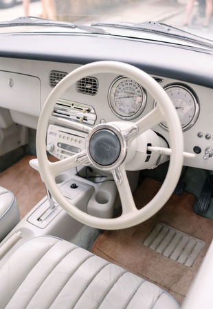 Cream vintage car interior with a white steering wheel, chrome-accented retro gauges and radio, quilted white leather seat and tan carpet floor mat.