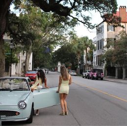 Two women in summer dresses by a pale blue vintage convertible parked on a tree-lined oak-and-palm street with historic townhouses