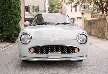 Front view of a mint-green vintage compact car with round headlights and honeycomb grille, parked on a quiet historic street with brick buildings and trees