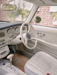 Right-hand-drive vintage car interior with cream leather seats and steering wheel, chrome round gauges, automatic shifter and beige carpet, brick house visible outside