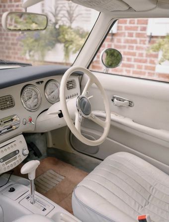 Right-hand-drive vintage car interior with cream leather seats and steering wheel, chrome round gauges, automatic shifter and beige carpet, brick house visible outside