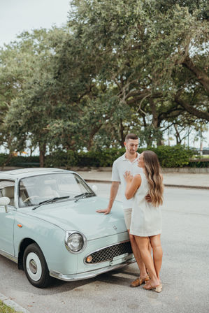 Smiling couple in light summer clothes leaning on a mint vintage car parked beneath sprawling oak trees on a sunlit tree-lined street