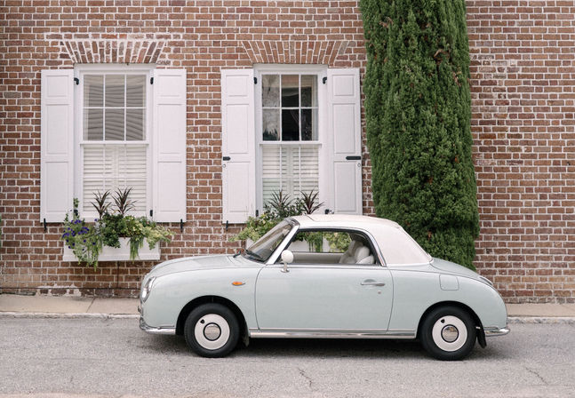 Charming pale mint retro compact car parked on a quiet street against a red brick facade with white shuttered windows, window boxes of greenery and a tall cypress tree