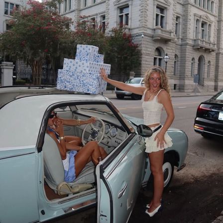 Two friends with stacked floral gift boxes on the roof of a light-blue vintage convertible parked on a historic downtown street in front of a stone building, one seated behind the wheel and one laughing in a white summer outfit.