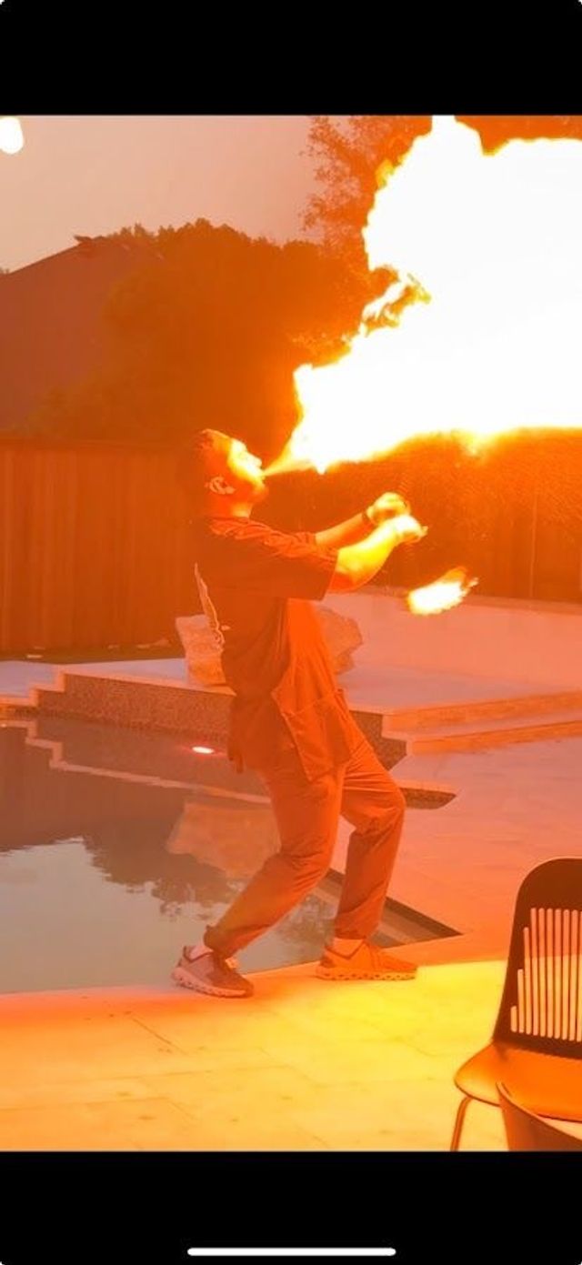 Fire performer breathing a huge plume of flame on a backyard pool deck at dusk, orange glow reflecting on the water and patio.