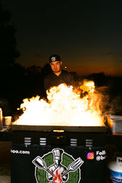 Outdoor chef in black uniform cooking over dramatic open flames on a flat-top grill at dusk, street-food style night market scene with sunset silhouette