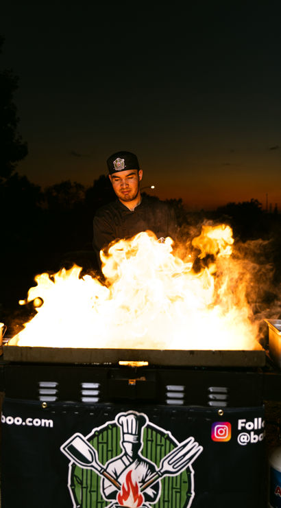 Outdoor chef in black uniform cooking over dramatic open flames on a flat-top grill at dusk, street-food style night market scene with sunset silhouette