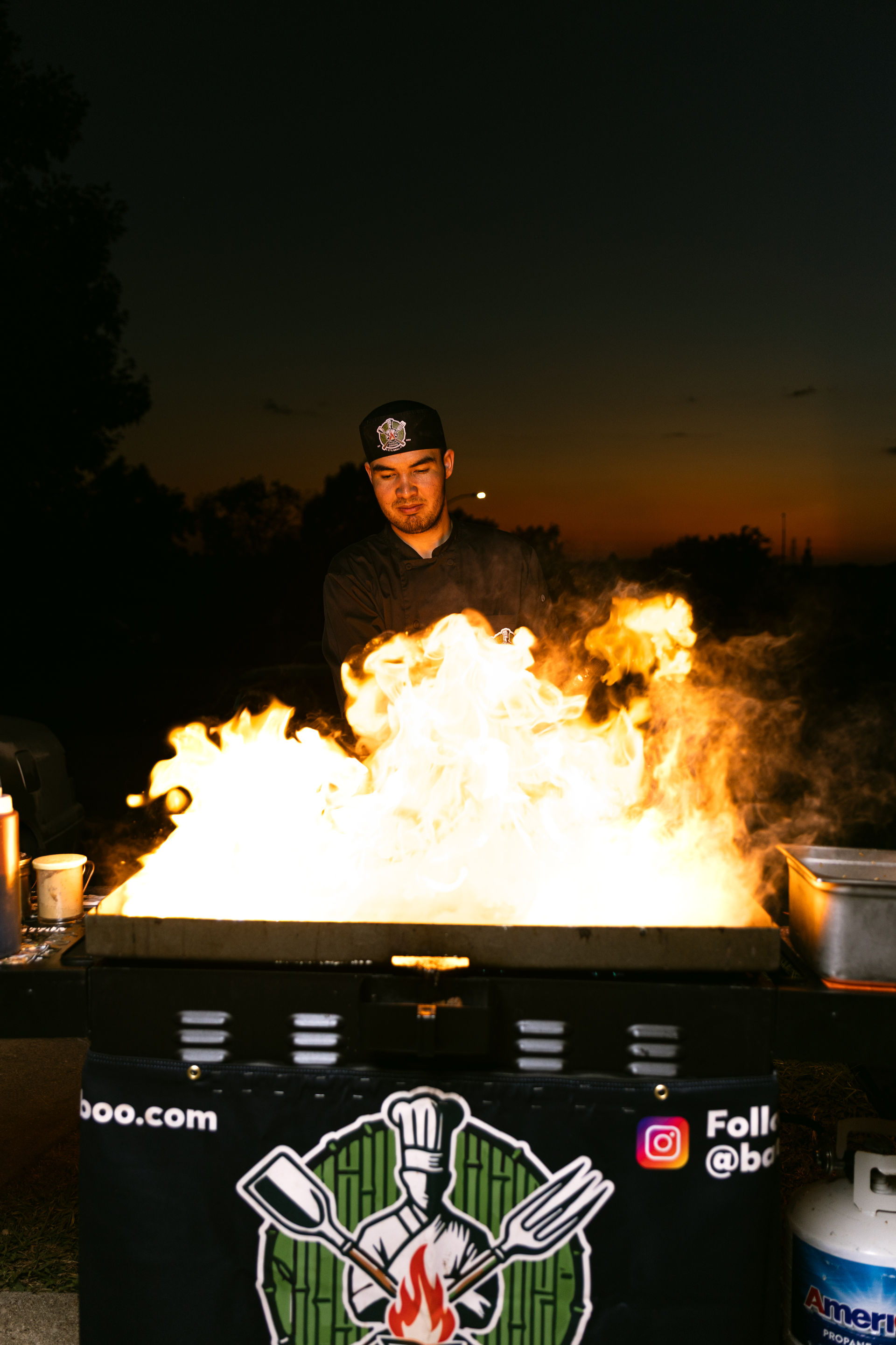 Outdoor chef in black uniform cooking over dramatic open flames on a flat-top grill at dusk, street-food style night market scene with sunset silhouette