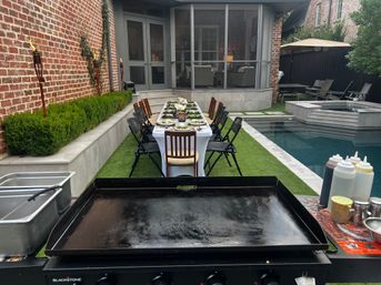 Poolside backyard patio set for al fresco dining — long linen-covered table with place settings, tiki torches, outdoor griddle and screened porch
