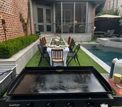 Poolside backyard patio set for al fresco dining — long linen-covered table with place settings, tiki torches, outdoor griddle and screened porch