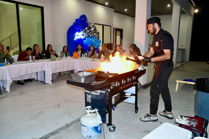Hibachi chef flambéing food on a flat-top griddle at an outdoor patio birthday party, guests seated at long tables watching and filming near blue balloon decor.