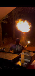 Backyard night scene of a person performing fire-breathing with a handheld torch over an outdoor grill, a large orange flame burst against dark trees.