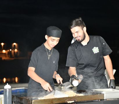 Two smiling cooks in black uniforms stir rice on a steaming flat-top grill at a nighttime poolside outdoor event with warm building lights reflecting on the water.