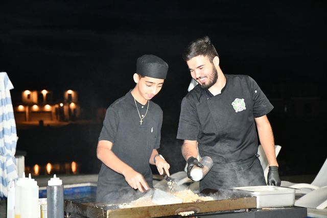 Two smiling cooks in black uniforms stir rice on a steaming flat-top grill at a nighttime poolside outdoor event with warm building lights reflecting on the water.