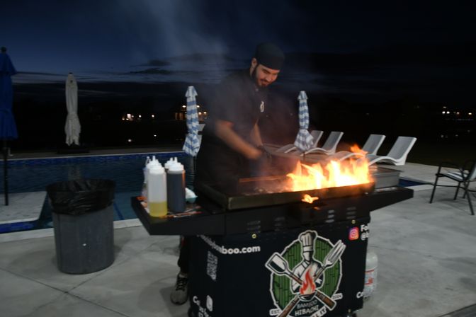 Poolside hibachi chef grilling over bright flames at night on an outdoor patio, mobile grill cart with condiment bottles, lounge chairs and umbrellas beside a swimming pool.