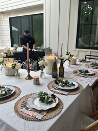 Cozy backyard patio dinner set for six with vintage plates of green salad, lit candles, wine bottles and a chef grilling in the background