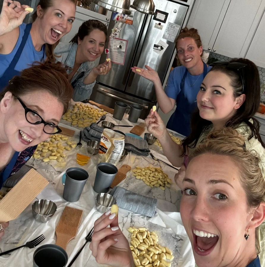 Six women smiling in a home kitchen making homemade gnocchi — holding small ridged potato dumplings over a marble countertop with gnocchi boards, trays of shaped pasta, cups and utensils.
