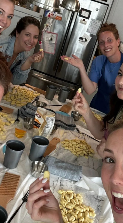 Six women smiling in a home kitchen making homemade gnocchi — holding small ridged potato dumplings over a marble countertop with gnocchi boards, trays of shaped pasta, cups and utensils.