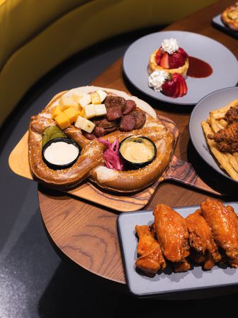 Shareable soft salted pretzel board with cubed cheeses, sliced sausages, pickles and two dipping sauces, served alongside buffalo chicken wings, fries and a strawberry dessert on a wooden restaurant table