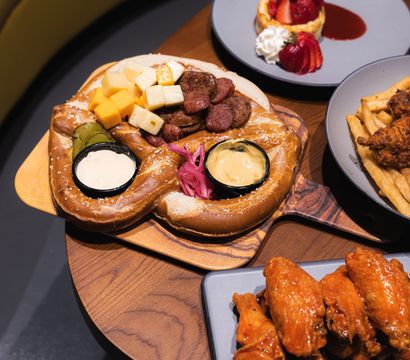 Shareable soft salted pretzel board with cubed cheeses, sliced sausages, pickles and two dipping sauces, served alongside buffalo chicken wings, fries and a strawberry dessert on a wooden restaurant table