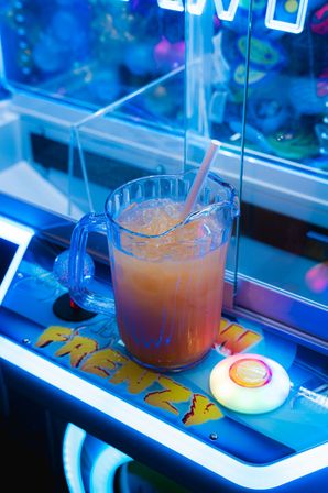Clear plastic pitcher filled with iced orange drink and a straw sitting on a neon-lit claw machine control panel in an arcade.