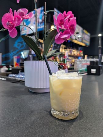 Iced creamy cocktail in a cut-glass tumbler with a black straw on a bar counter, bright pink orchid in a white ribbed pot and blurred bar shelves in the background.