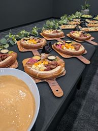 Pretzel grazing boards on wooden paddles at an indoor catered event — each topped with cheese cubes, sliced sausage, pickled onions and dipping sauces, with a large bowl of creamy dip and greenery decor.