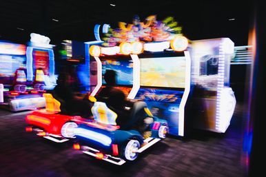 Neon-lit arcade racing game with two players in colorful car seats, motion-blurred action on large screens inside an indoor arcade entertainment center.