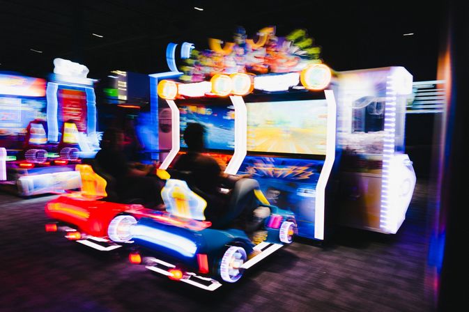 Neon-lit arcade racing game with two players in colorful car seats, motion-blurred action on large screens inside an indoor arcade entertainment center.
