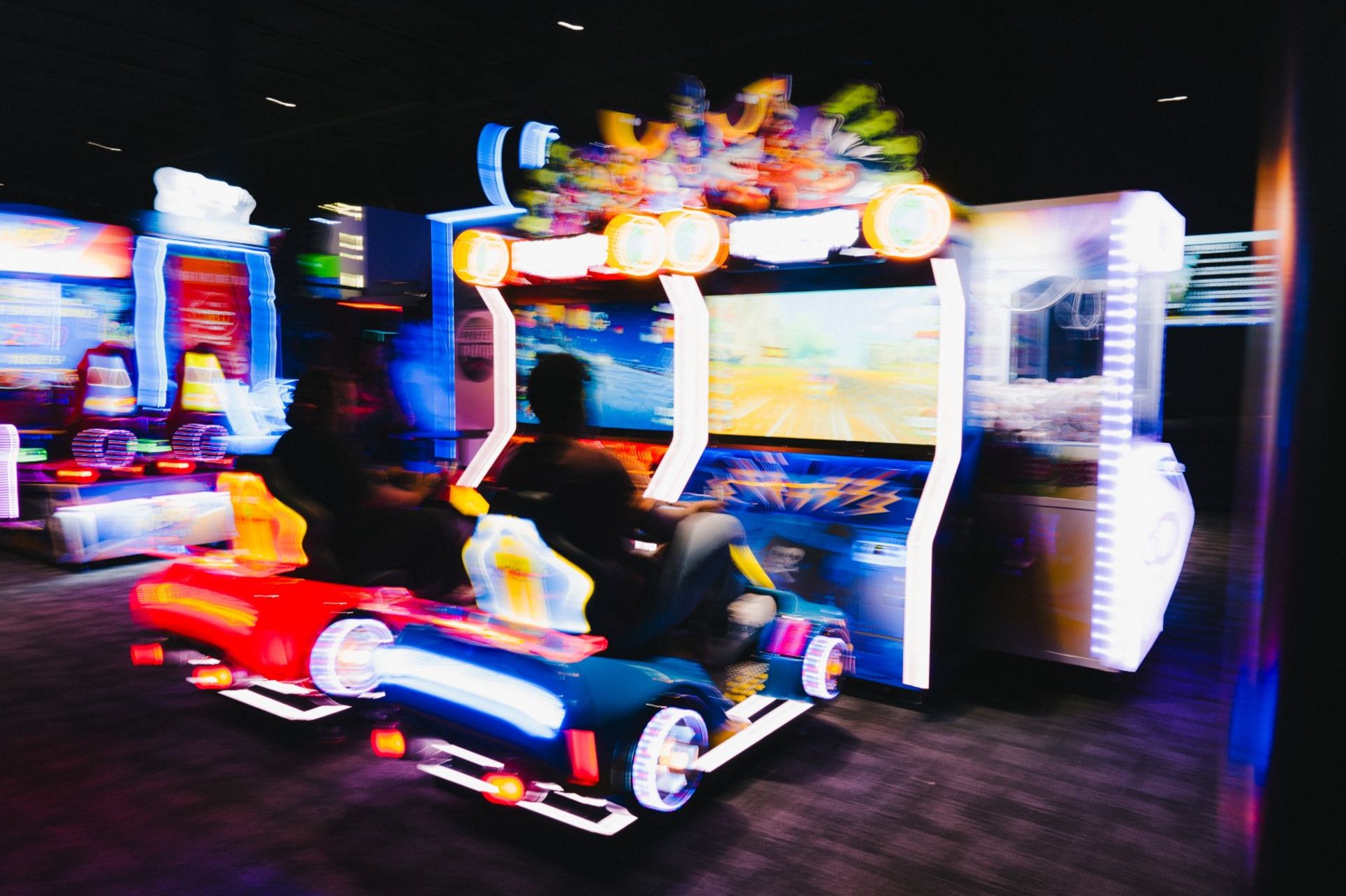 Neon-lit arcade racing game with two players in colorful car seats, motion-blurred action on large screens inside an indoor arcade entertainment center.