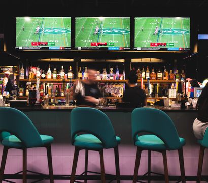 Vibrant sports bar scene with three teal bar stools at a counter, a stocked back bar of liquor bottles, blurred bartenders in motion, and three large TVs showing a football game.