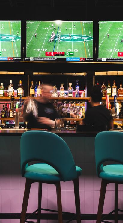 Vibrant sports bar scene with three teal bar stools at a counter, a stocked back bar of liquor bottles, blurred bartenders in motion, and three large TVs showing a football game.