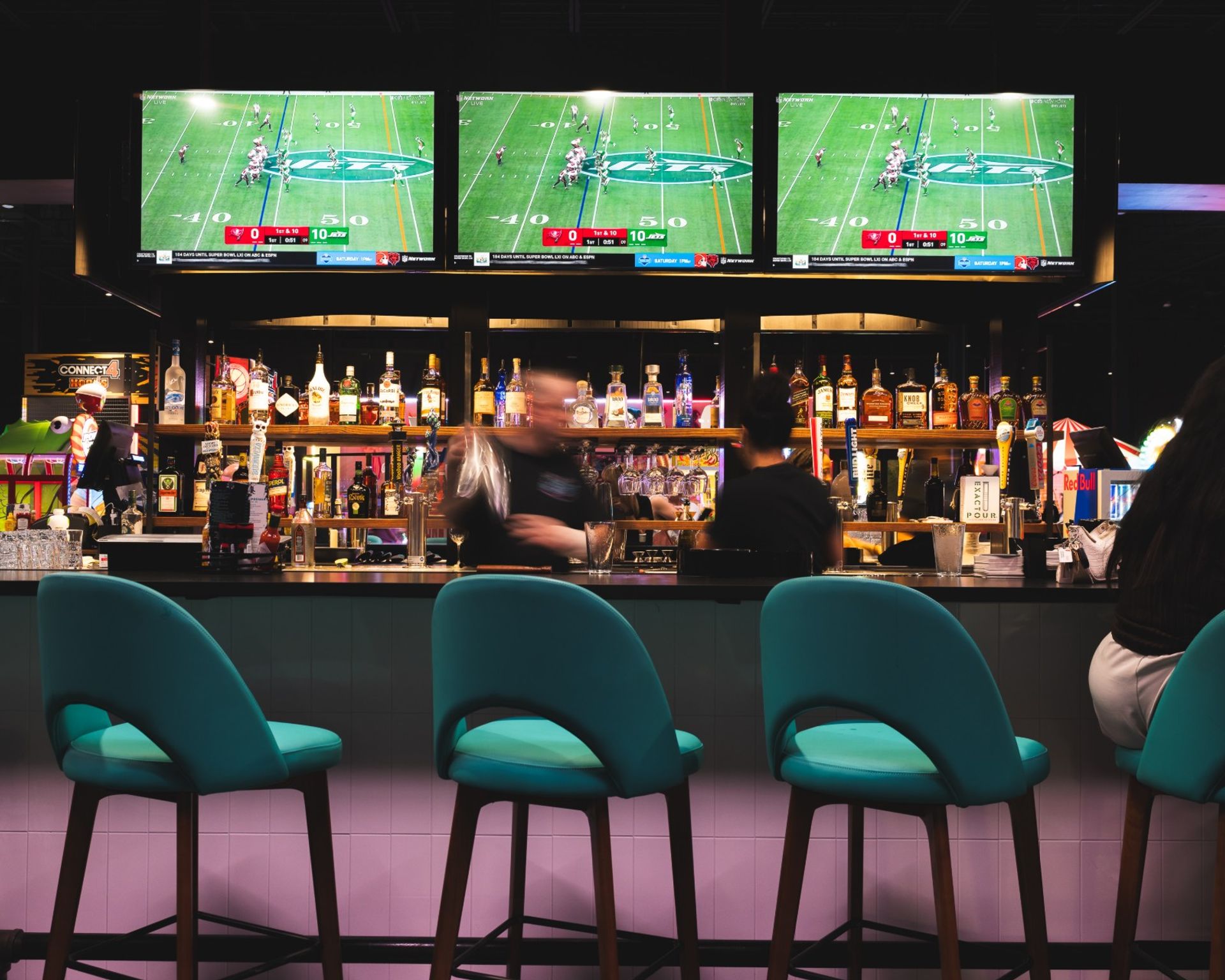 Vibrant sports bar scene with three teal bar stools at a counter, a stocked back bar of liquor bottles, blurred bartenders in motion, and three large TVs showing a football game.