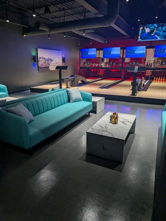 Modern bowling alley lounge with teal sofas, marble coffee tables, illuminated lanes and digital score screens in a stylish indoor entertainment space.