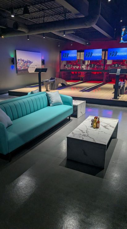 Modern bowling alley lounge with teal sofas, marble coffee tables, illuminated lanes and digital score screens in a stylish indoor entertainment space.
