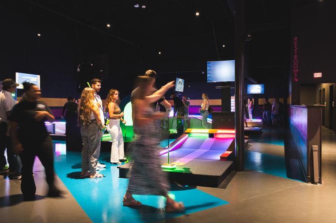 Neon-lit indoor mini golf with adults playing and mingling around colorful LED ramps and screens in a dim entertainment venue.