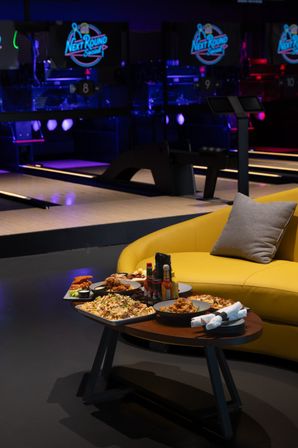 Neon-lit bowling alley lounge with lanes in the background, a yellow sofa and a low table stacked with wings, loaded fries, nachos and dipping sauces for a casual night out.