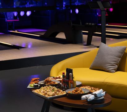 Neon-lit bowling alley lounge with lanes in the background, a yellow sofa and a low table stacked with wings, loaded fries, nachos and dipping sauces for a casual night out.