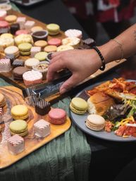Hand using tongs to grab a chocolate petit four from a wooden dessert board filled with colorful macarons and petit fours at a buffet, plate of slider, pasta and salad nearby
