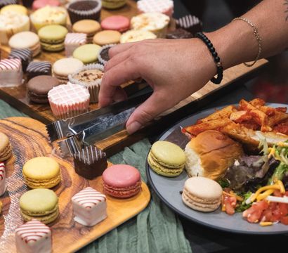 Hand using tongs to grab a chocolate petit four from a wooden dessert board filled with colorful macarons and petit fours at a buffet, plate of slider, pasta and salad nearby