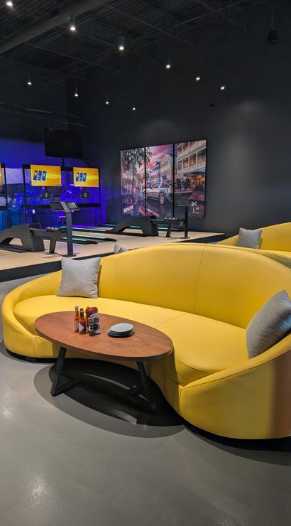 Vibrant yellow curved sofa and wooden coffee table in a modern indoor bowling alley lounge with neon-lit lanes, digital score screens and a mural in the background.