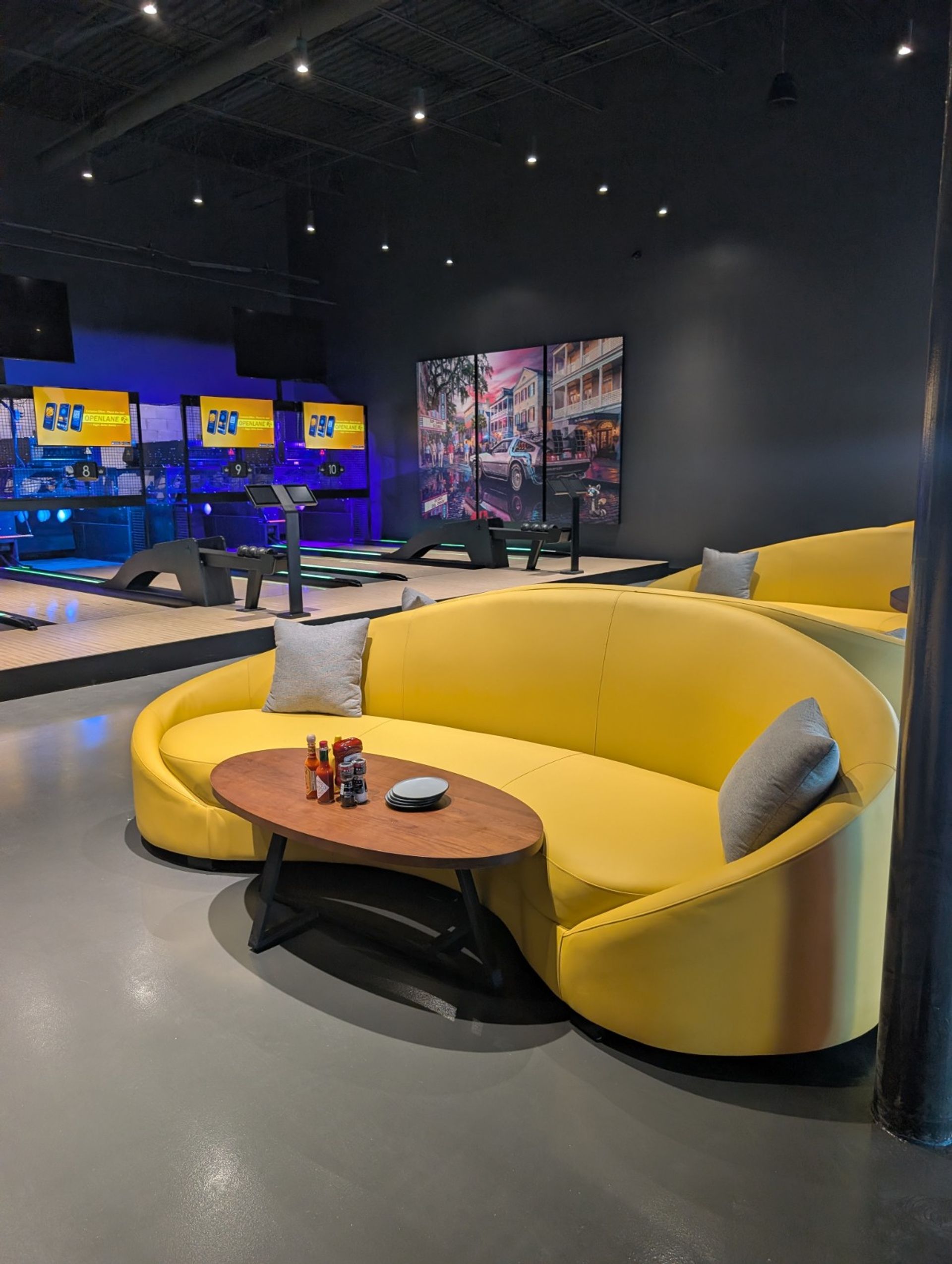 Vibrant yellow curved sofa and wooden coffee table in a modern indoor bowling alley lounge with neon-lit lanes, digital score screens and a mural in the background.